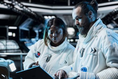 Takım çalışması yapan astronotlar sensörleri ayarlıyor, kıyafet bütünlüğünü doğrulamak için laptop kullanıyorlar. Uzay aracındaki kozmonotlar, rutin ekipman kontrolü sırasında ileri mühendislik teçhizatlarıyla çalışıyor.