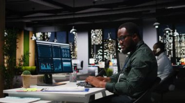 Modern çalışma alanındaki siyahi erkek yönetici bilgisayar monitöründeki tahminleri ve iş stratejilerini analiz ederek, gece geç saatlerde üretkenlik için rapor yazma ve planlama garantisi veriyor. Kamera A.