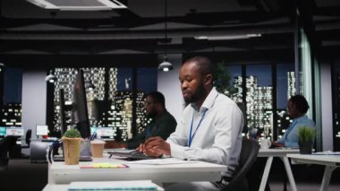 Afrika kökenli Amerikalı erkek işçi mali numaraları düzeltiyor, strateji odasında saatler sonra arşivlenmiş dosyaları inceliyor. Raporları değerlendiren ve araştırma yapan bir adam. Kamera A.