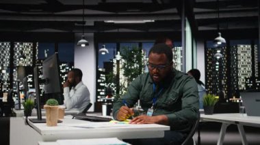 Siyahi iş adamı monitöre yapışkan bir not bırakıyor. Yeni bir gelişim görevini hatırlamak için yapışkan kağıt etiketi kullanıyor. Geceleri karanlık ofis, verimlilik motivasyonu. Kamera A.