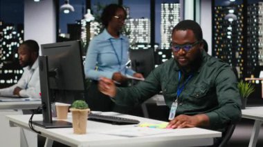 Siyahi iş adamı monitöre yapışkan bir not bırakıyor. Yeni bir gelişim görevini hatırlamak için yapışkan kağıt etiketi kullanıyor. Geceleri karanlık ofis, verimlilik motivasyonu. Kamera B.