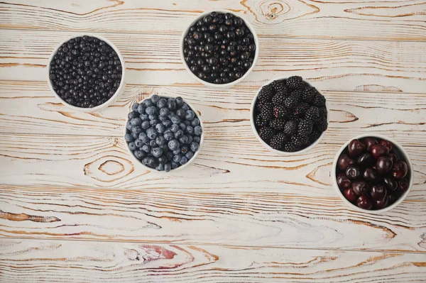 five bowls with seasonal berries, a mix of fresh berries on a wooden table, gooseberries, blueberries, currants, cherries, blueberries, blackberries,