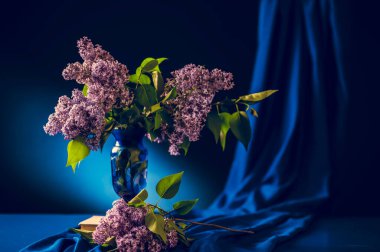 Masadaki vazodaki leylak çiçekleri pencere kenarındaki bir atölyede mavi bir arka plana karşı.