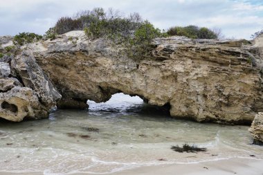 A unique rock formation features a natural arch, surrounded by water and coastal vegetation. The cloudy sky adds a serene atmosphere to this peaceful beach setting. Sandy Cape Jurien Bay Western Australia clipart