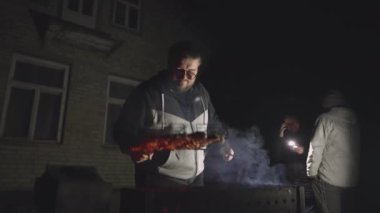 A young man cooks meat on the grill against the backdrop of a village house and two guys talking to each other