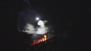 A young man with a flashlight in his hand cooks soup in a cauldron on a brazier