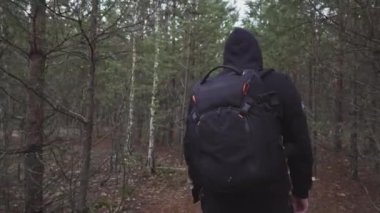 A young man in a hoodie and with a backpack, walking through the forest, stops to look around.