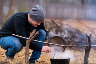 A young man prepares soup in a cauldron suspended over a fire, stirring it with a large spoon.
