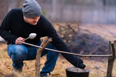 The guy holds a large spoon in one hand, and with the other he lifts the lid of the cauldron hanging over the fire.