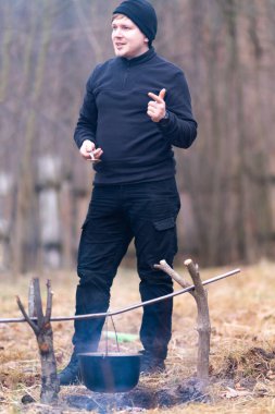 The guy, holding a large spoon in his hand, stands by the fire with cooked food, and mentally lists the ingredients that he put into the cauldron.