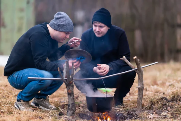 The guys cook food in a cauldron over a fire and one of them tastes the broth