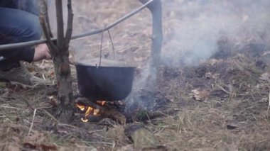 A hiker in the forest throws tree branches into the fire to cook food in a cauldron.