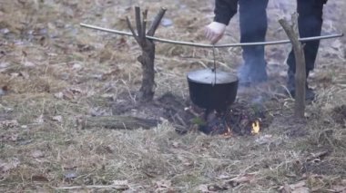 A man prepares food in a cast-iron cauldron that hangs over an open fire.
