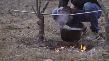 A young man stirs food with a large spoon in a cauldron hanging over a fire.