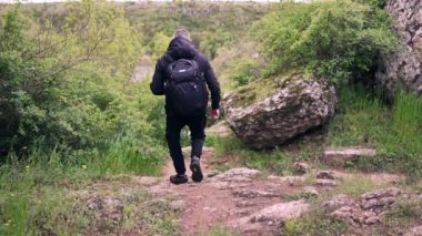 Siyah elbiseli bir adam ve sırt çantalı bir adam bir dağ yolu boyunca nehire iniyor..