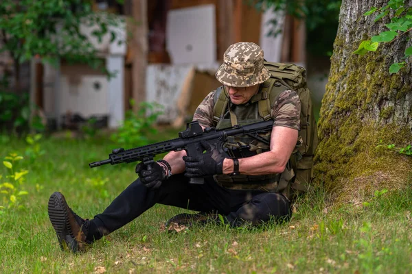 A man in a camouflage uniform, sitting under a tree, reloads a rifle