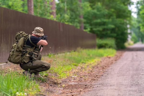 A warrior watches the road through the scope of his rifle, crouching on the side of a dirt road.