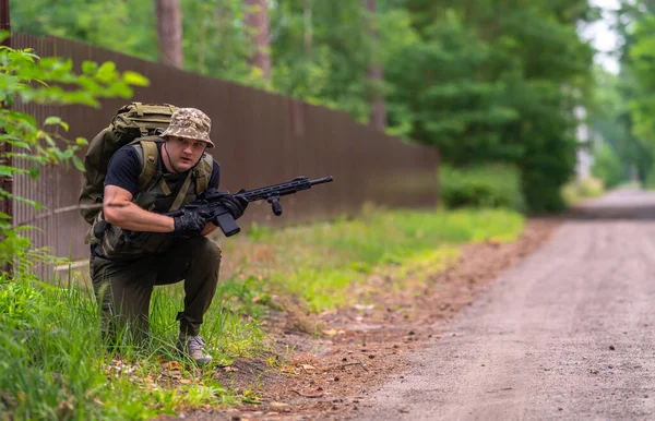 バックパックとライフルを持った軽くて軍事的な制服を着た男が茂みの後ろから覗く — ストック写真