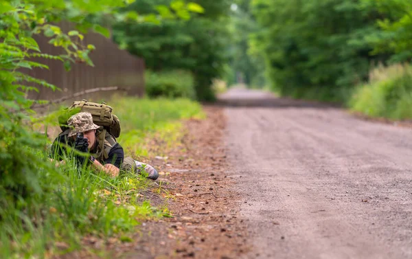 軍の制服を着た男が道の脇に横たわっている間ライフルの範囲を調べる — ストック写真