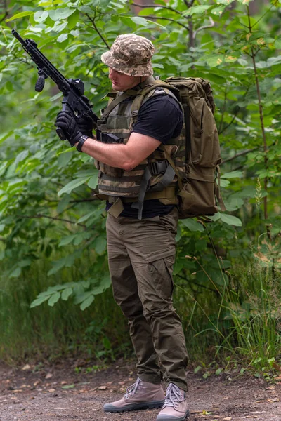 軍の制服を着た男が森の脇に立っている間に自動小銃を装填する — ストック写真
