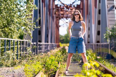 Kız demiryolu raylarının üzerinde duruyor metal yapılar köprüsünden geçiyor.