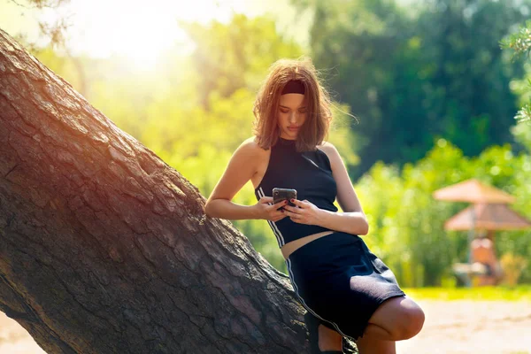 Девушка Фоне Заходящего Солнца Глядя Телефон Прислонившись Дереву Лицензионные Стоковые Изображения