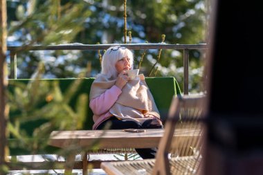 Yetişkin bir kadın, battaniyeyle kaplı bir sandalyede otururken elinde bir bardak tutuyor ve omuzlarına atkı atıyor..