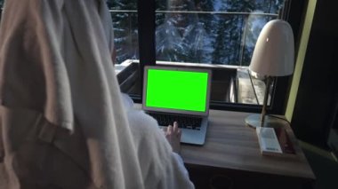 Spa tedavisinden sonra bornozlu bir kadın pencerenin dışındaki kış ormanının arka planına karşı masasında dizüstü bilgisayar kullanıyor. Yeşil Ekran.
