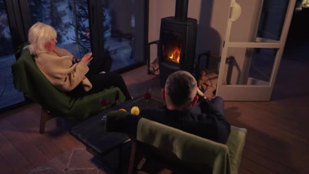 一对男女在他们的手机上分享照片 而舒适地坐在壁炉前的扶手椅上 — 图库视频影像