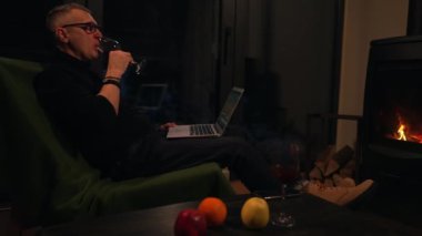 Yetişkin bir adam, bir sandalyede oturup kırmızı şarap içerken dizüstü bilgisayardan internet içeriğini izliyor..