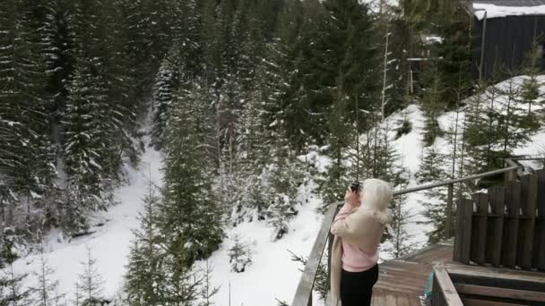 女性は彼女の山のシャレーのテラスに立っている間 冬の森の写真を撮ります — ストック動画