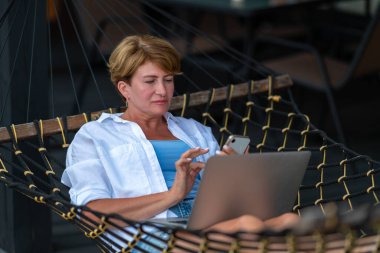 Hamakta yatarken kadın bir telefon ve dizüstü bilgisayar kullanıyor..