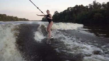 Siyah mayo giymiş bir kız wakeboard yapıyor, tek eliyle bir tekneye bağlı bir halata tutunuyor..