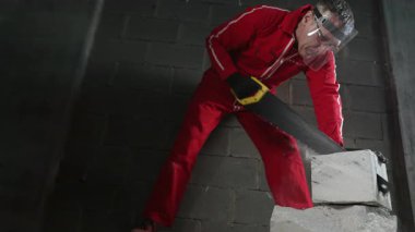 Koruyucu takım elbiseli ve maskeli bir adam tuğlaları el testeresiyle parçalara ayırıyor..