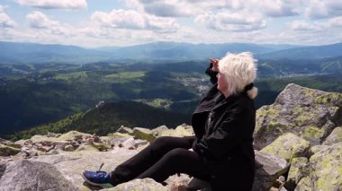 Orta yaşlı bir kadın, bir dağın tepesine tırmanmış, elini alnına koyup uzaklığa bakar..