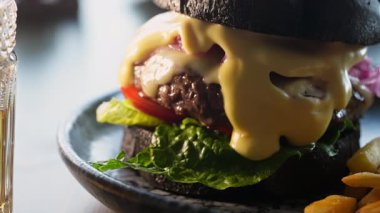 Biftek ve peynirli hamburgerin siyah bir çöreğin içinde bir bardak biranın yanında..