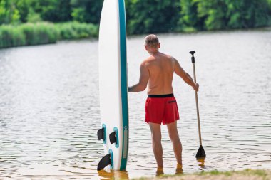 Bir adam bir nehir veya gölün kumlu kıyısında duruyor, küreği ve SUP tahtasıyla..