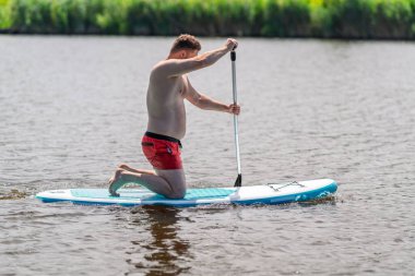 Bir adam nehir ya da göl kenarındaki bir SUP tahtasının üzerinde yüzerken yaz güneşinin tadını çıkarıyor..