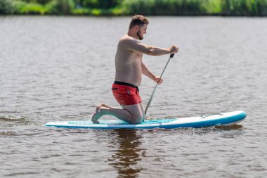 Bir adam yaz günü nehir ya da göl kenarında SUP tahtasının üzerinde yüzüyor..