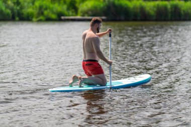 Sıcak bir yaz günü nehir ya da göl kıyısında SUP tahtasının üzerinde yüzen bir adam var..