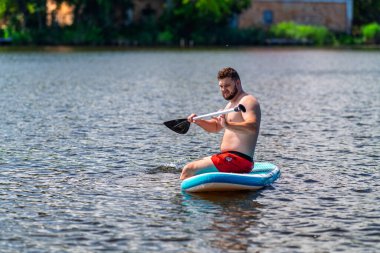 Sakallı genç bir adam nehrin ortasında bir SUP tahtasının üzerinde otururken eline bir kürek alır..