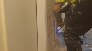Sarı yüksek görüş yeleği giyen sıva iç duvardan eski alçıyı kaldırıyor, çok toz yapıyor, el kamerasıyla ateş ediyor..