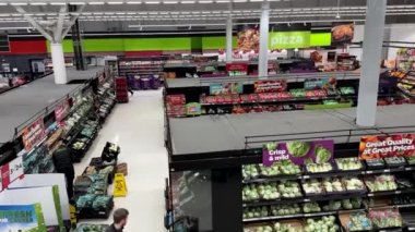 Asda süpermarketinin içinde yürüyen merdivenden aşağıya doğru yüksek açılı görüntü. Alışveriş ve tezgahtarlar sebze ve meyve stokluyor.