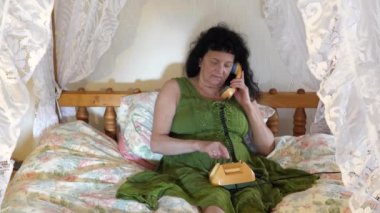 Uzun siyah kıvırcık saçlı, yeşil retro tarzı elbisesi olan orta yaşlı bir kadın. Paraşütlü yatakta telefon numarası çeviriyor. Konuşurken gülümsüyor ve antika telefonu kaldırıyor..