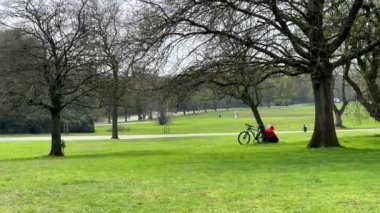 Şehir parkında ilkbahar manzarası. Manchester, İngiltere 'de Heaton Park' ta ağaçta oturan bisikletli bir adam, yanından geçen bir aile. Tanınmayan insanlar.