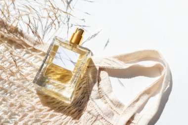 Transparent bottle of perfume with label and cloth shopping bag, wildf grass on white background. Fragrance presentation with daylight. Trending concept in natural materials. Womens and mens essence