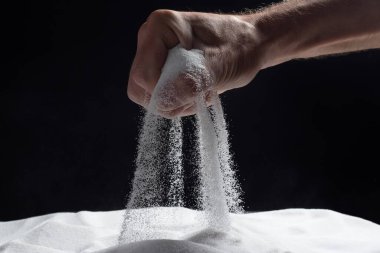Elinde bir avuç dolusu kuru kum olan bir adam, siyah arka planda parmaklarının arasından kumu döküyor. Yaşam akışı kavramı. Saf doğal mineral kuvars tanelerini kapatın..