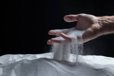 Elinde bir avuç dolusu kuru kum olan bir adam, siyah arka planda parmaklarının arasından kumu döküyor. Yaşam akışı kavramı. Saf doğal mineral kuvars tanelerini kapatın..