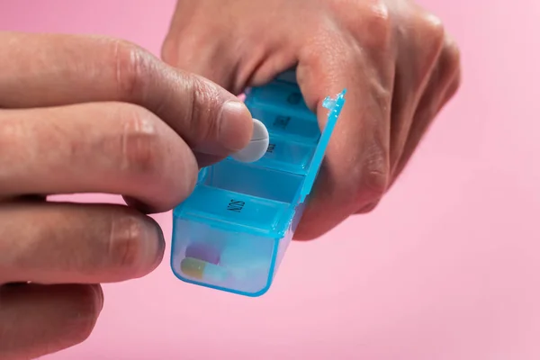 Man take pills from box. Pill organizer with variety of pills and supplement in hands. Weekly Pill Organizer on pink background. Daily pill box with medications and nutritional supplements.
