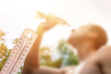 Aşırı sıcakta su içen adam, yaz gününde termometre, arka planda güneş olan yüksek sıcaklığı gösterir veya gösterir..